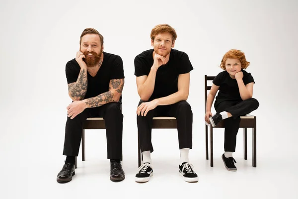 Full length of redhead kid with bearded grandpa and dad sitting on chairs with hands near face on grey background — Stock Photo
