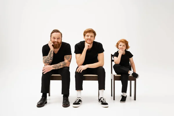 Full length of happy redhead kid with bearded grandfather and dad sitting on chairs and smiling at camera on grey background — Stock Photo