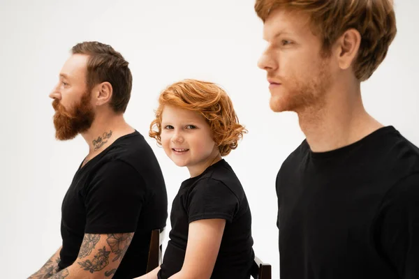 Cheerful redhead boy looking at camera near bearded tattooed grandfather and dad isolated on grey — Stock Photo