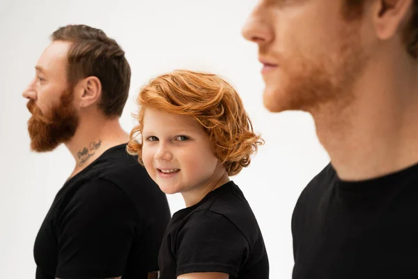 Happy red haired boy smiling at camera near bearded tattooed grandpa and dad on blurred foreground isolated on grey — Stock Photo