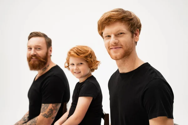Smiling man with redhead son and bearded tattooed father sitting and looking at camera isolated on grey — Stock Photo