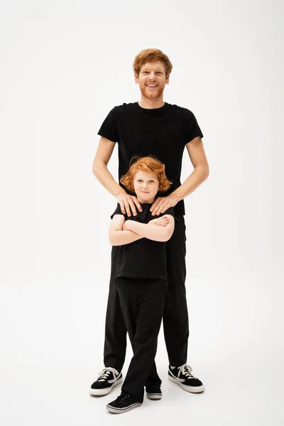 Full length of happy man hugging shoulders of red haired son standing with crossed arms on light grey background — Stock Photo