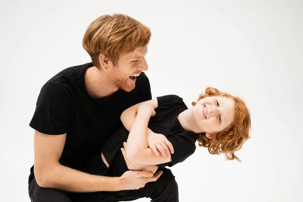 Excited man in black t-shirt playing with happy redhead son crossing arms isolated on grey — Stock Photo
