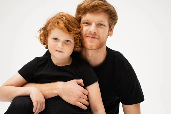 Happy redhead man hugging smiling son while looking at camera isolated on grey — Stock Photo