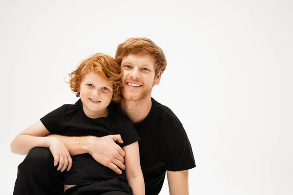 Pleased man in black t-shirt embracing red haired kid and looking at camera isolated on grey — Stock Photo