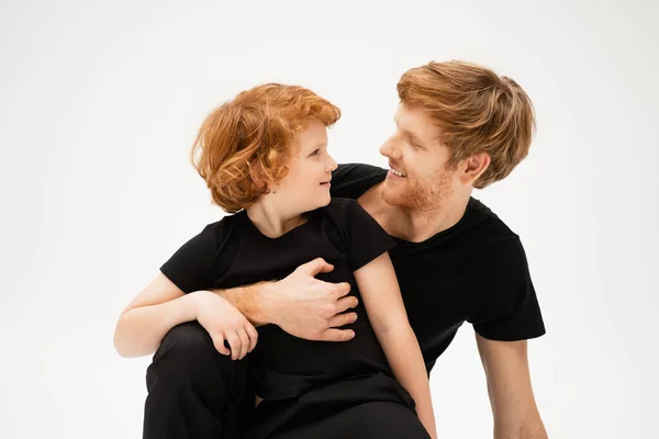 Happy man in black t-shirt embracing redhead son while looking at each other isolated on grey — Stock Photo