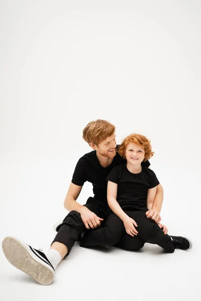 Full length of cheerful redhead boy smiling at camera while sitting with young father on light grey background — Stock Photo