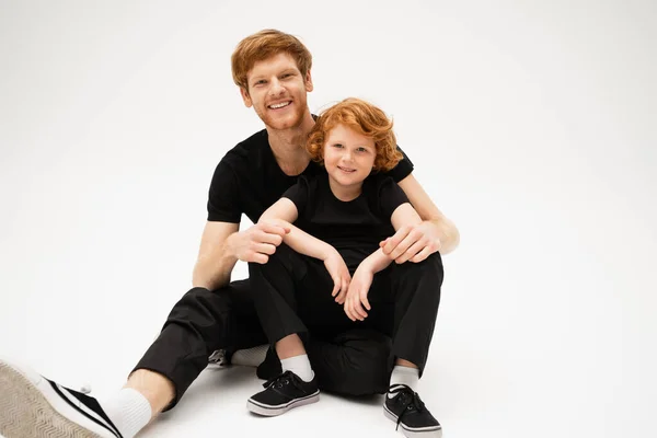 Joyful father and son with red hair smiling at camera while sitting on light grey background — Stock Photo