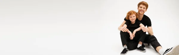 Joyful red haired dad and son in black clothes smiling at camera while sitting on light grey background, banner — Stock Photo