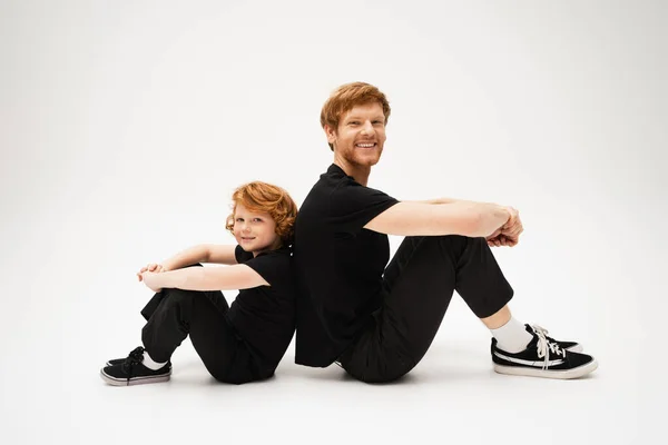Full length of happy red haired dad and son in black t-shirts and pants sitting back to back on light grey background — Stock Photo