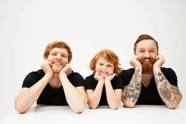 Happy boy with redhead father and bearded tattooed grandpa lying and smiling at camera on light grey background — Stock Photo