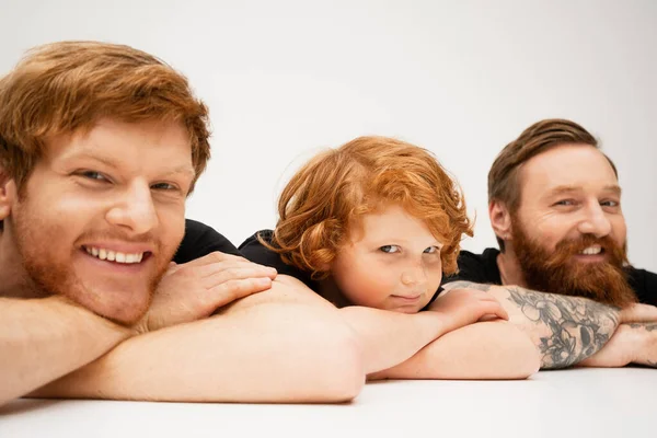Joyful man with redhead son and bearded tattooed dad lying with crossed arms and smiling at camera on light grey background — Stock Photo