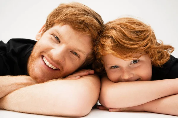 Family portrait of redhead father and son lying with crossed arms and smiling at camera on light grey background — Stock Photo