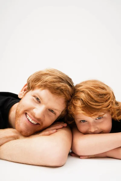 Young and cheerful man with red haired son lying with crossed arms and looking at camera on light grey background — Stock Photo
