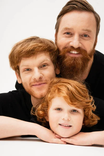 Family portrait of redhead kid with dad and bearded grandfather smiling at camera on light grey background — Stock Photo