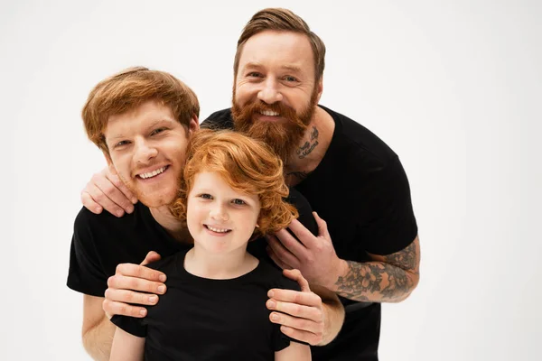 Family portrait of redhead men and cheerful boy in black t-shirts hugging and smiling at camera isolated on grey — Foto stock
