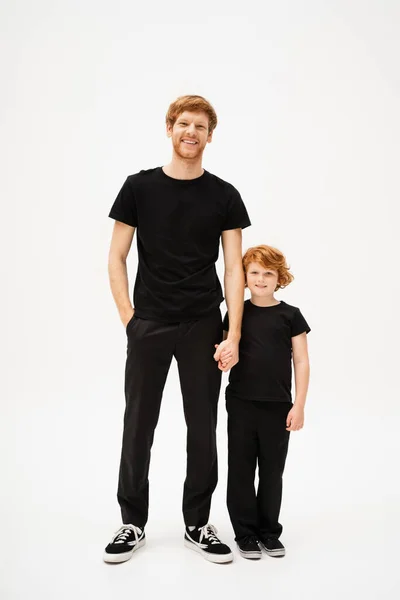 Full length of cheerful red haired dad and son holding hands and looking at camera while standing on light grey background — Stock Photo
