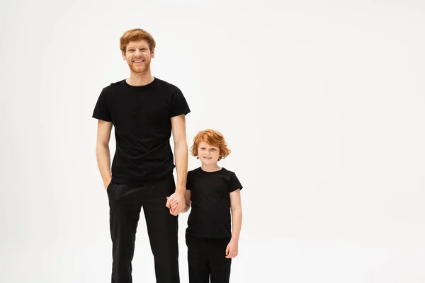 Joyful redhead father and son in black t-shirts and trousers holding hands and smiling at camera isolated on grey — Stockfoto