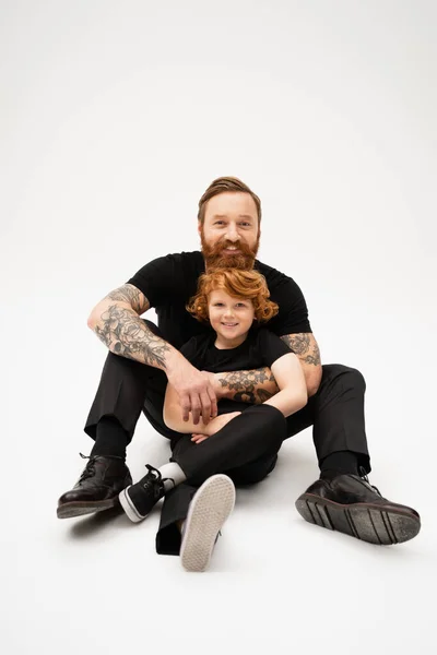 Full length of tattooed man with red haired grandson sitting in black clothes on light grey background — Stock Photo