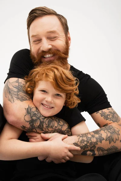 Cheerful tattooed man with closed eyes hugging smiling redhead grandson isolated on grey — Stock Photo