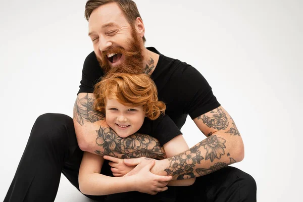 Cheerful tattooed man with closed eyes hugging happy redhead grandson on grey background — Stock Photo