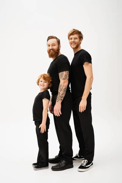 Full length of bearded redhead men and kid in black t-shirts and trousers smiling at camera while standing on light grey background — Stock Photo