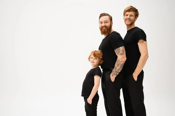 Smiling red haired men and kid in black t-shirts posing with hands in pockets of black trousers isolated on grey — Stock Photo