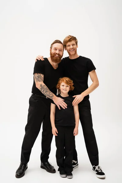 Full length of joyful bearded men and redhead kid in black t-shirts and trousers posing on light grey background — Stock Photo