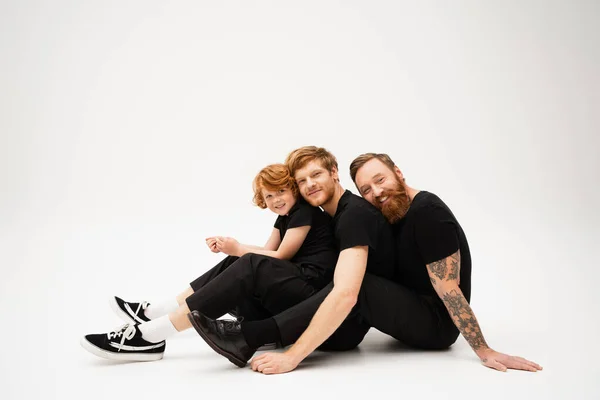 Joyful redhead men and boy in black clothes looking at camera while sitting on light grey background — Stock Photo