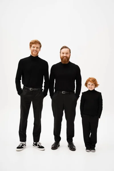 Full length of cheerful bearded men with red haired kid in black clothes standing with hands in pockets on light grey background — Stock Photo