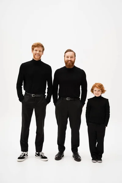 Full length of cheerful bearded men with red haired kid in black clothes standing with hands in pockets on light grey background — Stockfoto