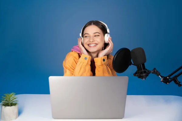 Cheerful teenager in headphones sitting near laptop and microphone isolated on blue - foto de stock
