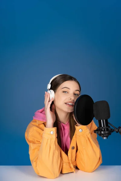 Brunette teenager touching headphones and talking near studio microphone isolated on blue — Foto stock