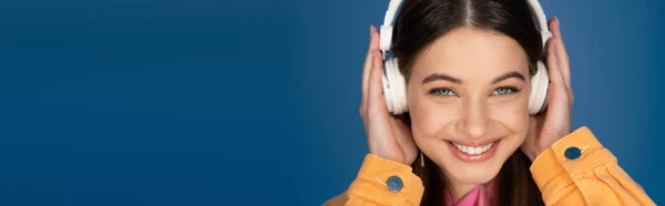 Positive teen girl in wireless headphones looking at camera isolated on blue, banner — Stock Photo