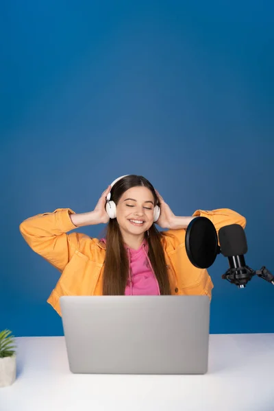 Smiling teen girl in headphones using laptop near microphone isolated on blue — Stock Photo
