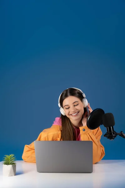 Teen podcaster in headphones looking at laptop near studio microphone isolated on blue - foto de stock