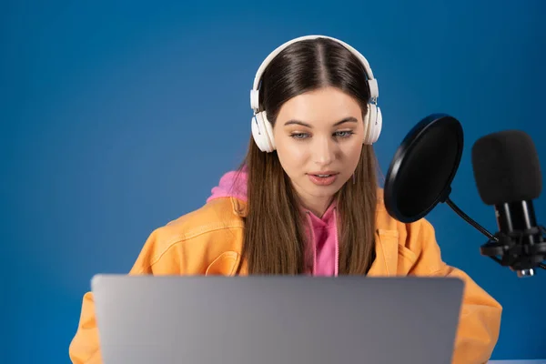 Teen podcaster in headphones looking at blurred laptop near studio microphone isolated on blue — Stock Photo