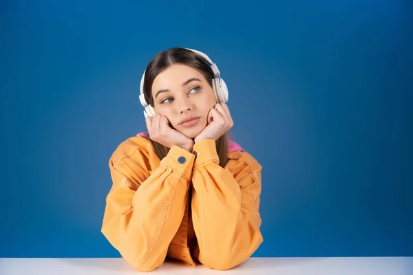 Dreamy brunette teen girl in headphones looking away near table isolated on blue — Stock Photo