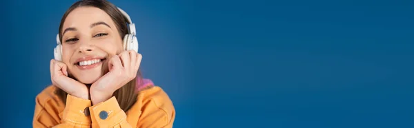 Positive teenage girl in headphones looking at camera isolated on blue, banner — Stock Photo