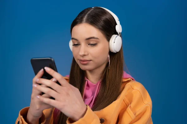 Teen girl in wireless headphones chatting on smartphone isolated on blue — Stock Photo