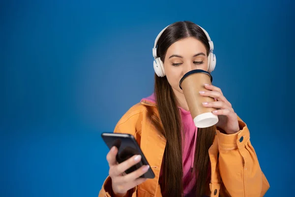 Teenage girl in headphones chatting on smartphone and drinking coffee to go isolated on blue - foto de stock