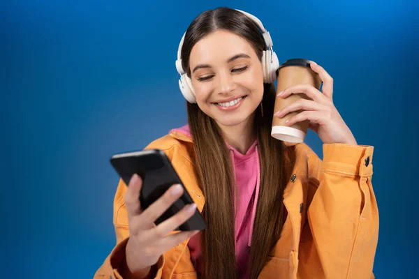 Positive teenager in headphones using cellphone and holding coffee to go isolated on blue — Stock Photo