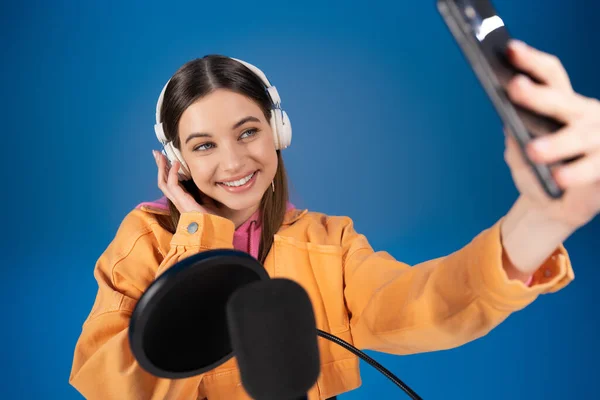 Teenager in headphones taking selfie on smartphone near blurred podcast microphone isolated on blue — Stock Photo