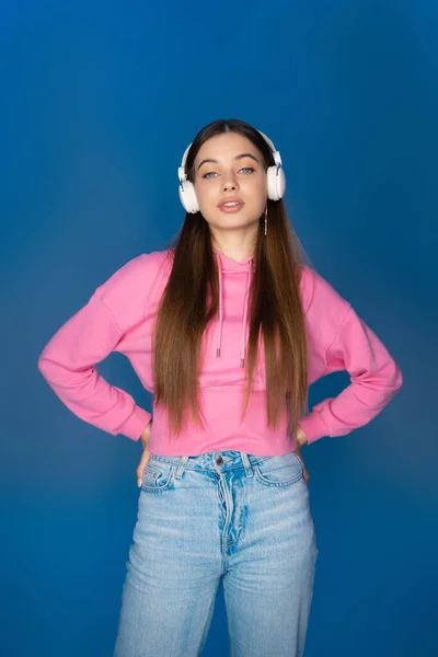 Teen girl in jeans and hoodie listening music in headphones isolated on blue - foto de stock