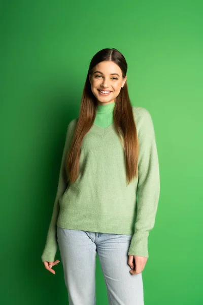Позитивный подросток в джинсах и свитере смотрит в камеру на зеленом фоне — стоковое фото