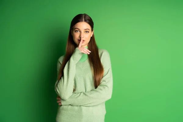 Brunette teen girl in jumper showing secret gesture on green background - foto de stock