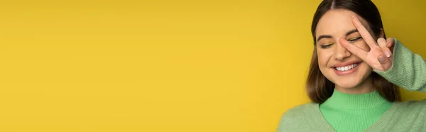 Cheerful brunette teen girl showing peace gesture on yellow background, banner — Foto stock