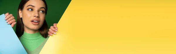 Teenager in jumper touching colorful paper and looking away isolated on green, banner — Stock Photo