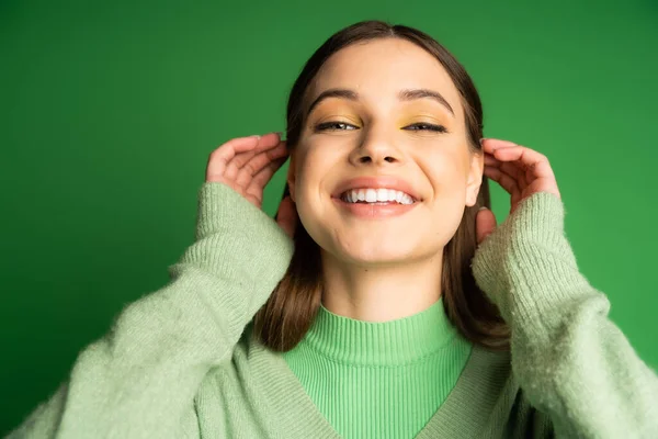 Cheerful teenager in cozy jumper adjusting hair isolated on green — Stock Photo