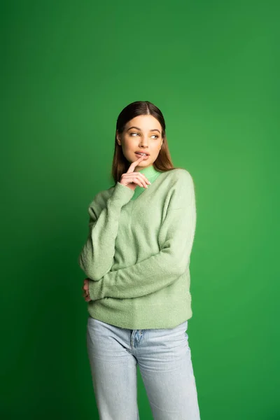 Pretty teen girl in jeans and warm jumper touching lip on green background — Stock Photo